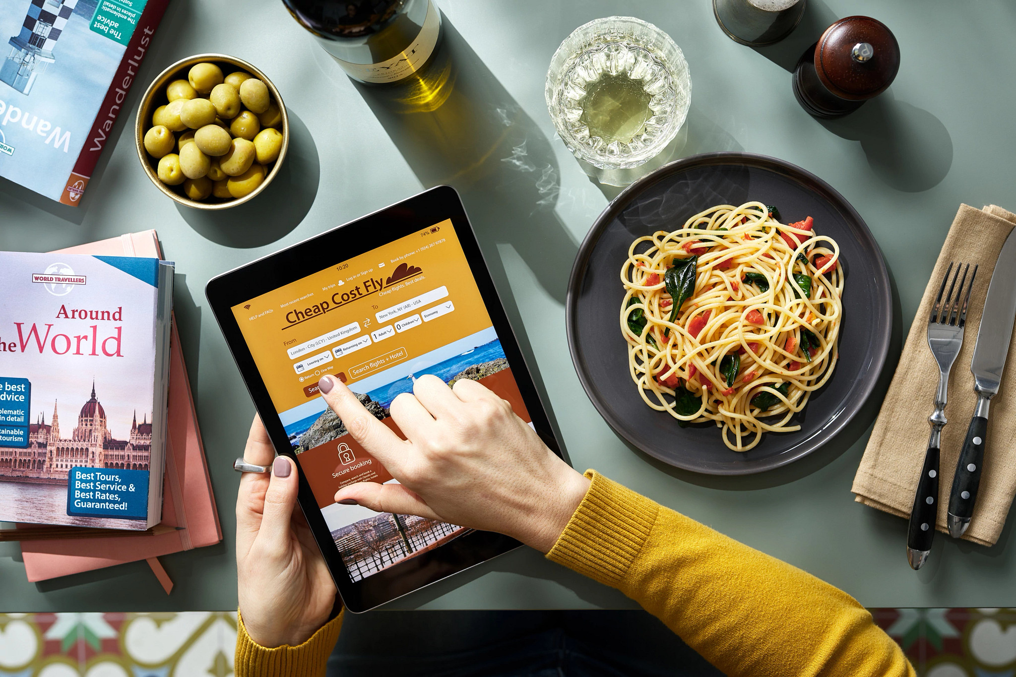 a person holding a tablet on a table with food, wine and travel books