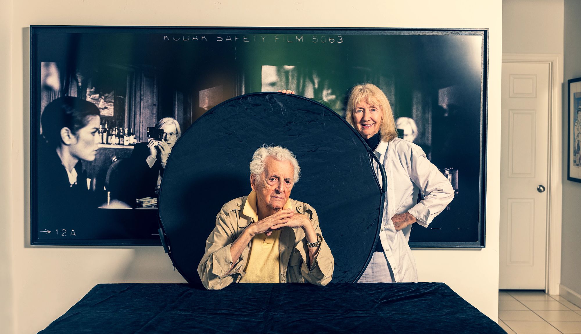 Harry Benson on President Nixon’s Resignation harry benson sits at a table. his wife stands next to him, and behind them is a large piece of wall art