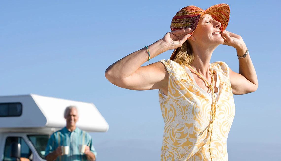 Mature woman with hands on her hat by a motor home on beach with her husband in the background. 