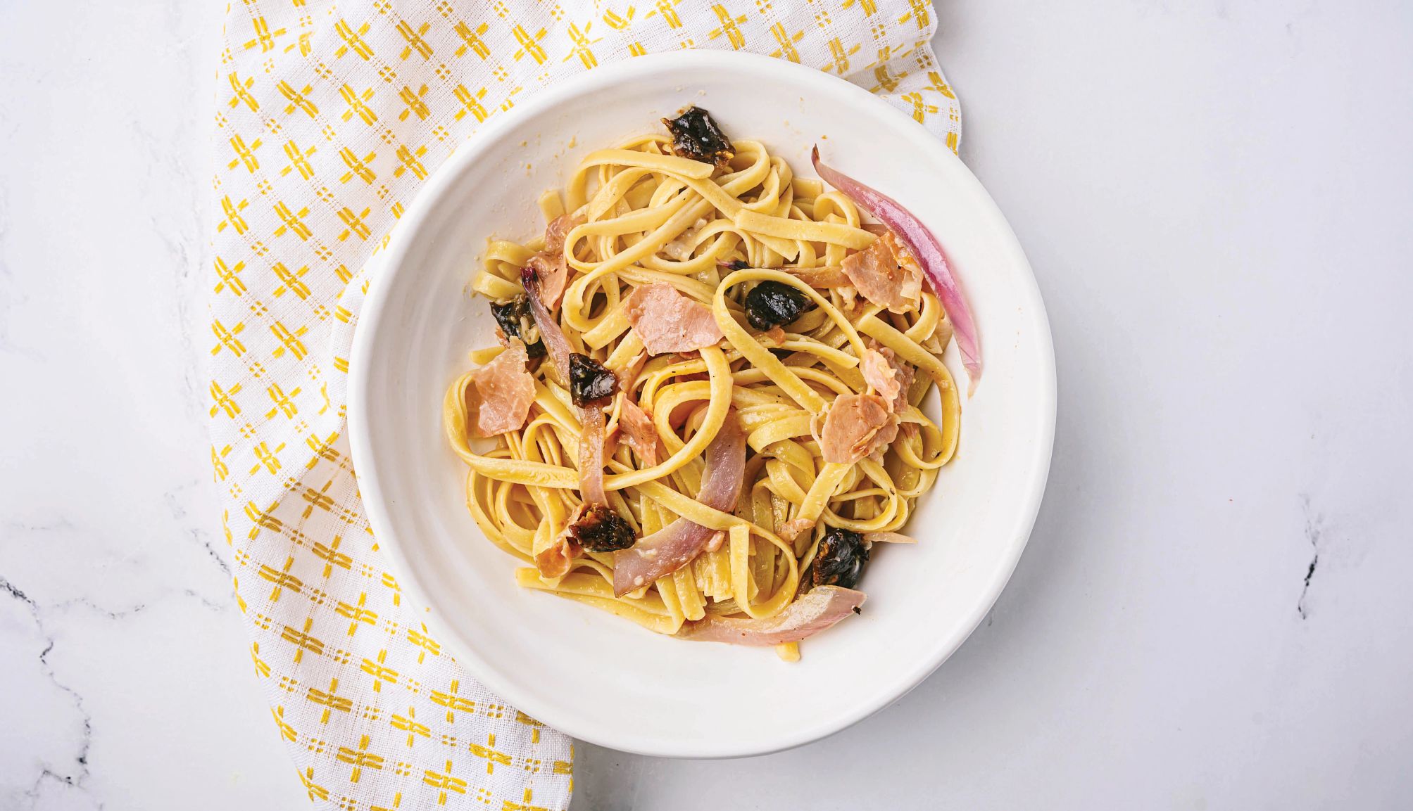 a bowl of fettuccine with prunes on a yellow and white patterned napkin on a table