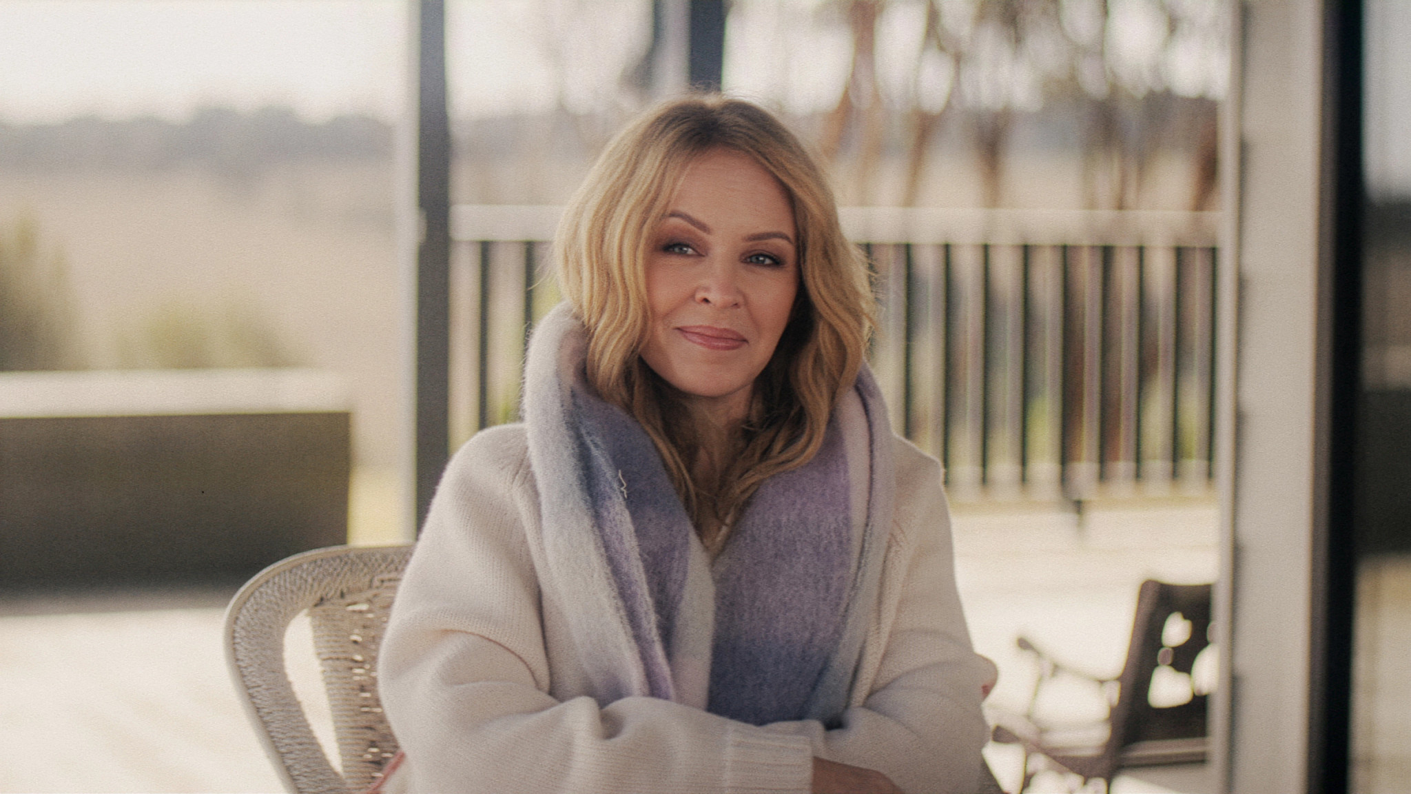 kylie minogue seated outdoors, wearing a sweater and scarf