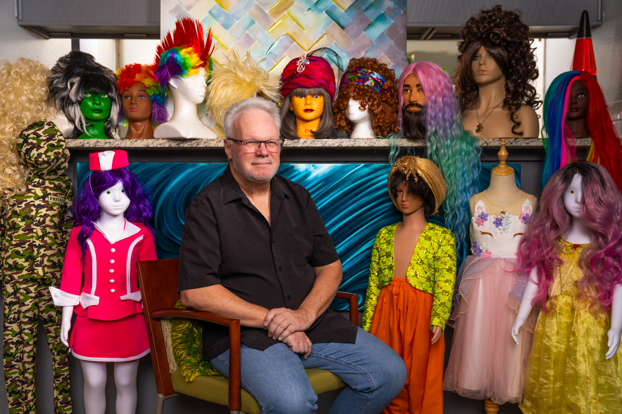 Christophe Waggoner is seated among many of his costumes. Some are on mannequins. There is a shelf of wigs above.