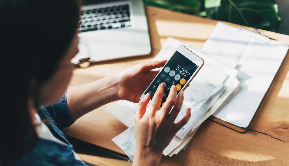 Woman using calculator on her phone while sitting at desk with laptop and financial documents on top