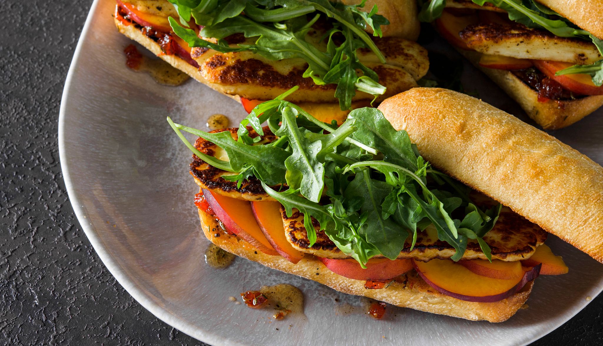 Fried Halloumi Sandwich A fried halloumi sandwich with peaches, arugula and honey is pictured on a plate atop a countertop.