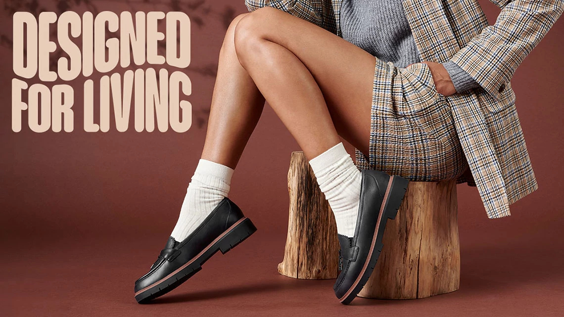 Clarks woman sitting, only showing lower part of body, wearing a plaid jacket and shorts suite with grey sweater, white socks, bare legs, black loafers, brown background, Designed for Living words in corner
