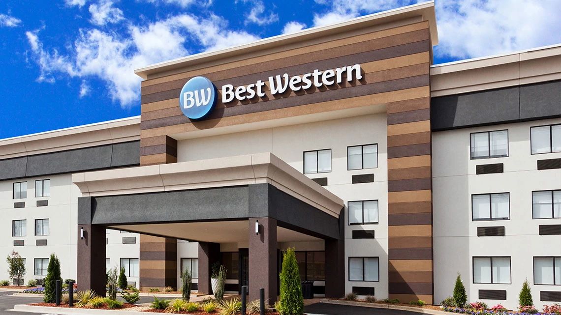 Best Western® front of a Best Western hotel with brown shades of striped siding, large covered area at the front entrance, parking lot, blue sky with white clouds