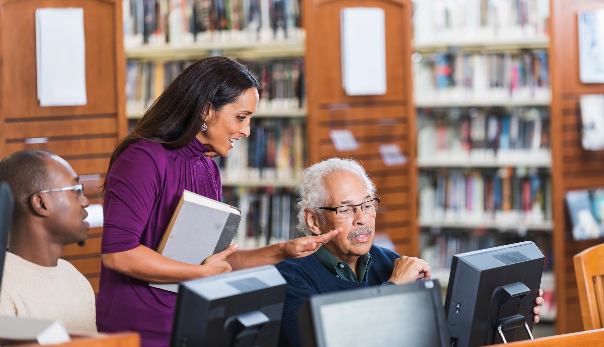 Senior Hispanic man in his late 70s using a computer in the library, getting help from a female librarian. He is sitting down, looking at the screen. She is standing beside him, talking and gesturing with her hand. A young African American man is watching and listening to their conversation. Senior Hispanic man in his late 70s using a computer in the library, getting help from a female librarian. He is sitting down, looking at the screen. She is standing beside him, talking and gesturing with her hand. A young African American man is watching and listening to their conversation.
