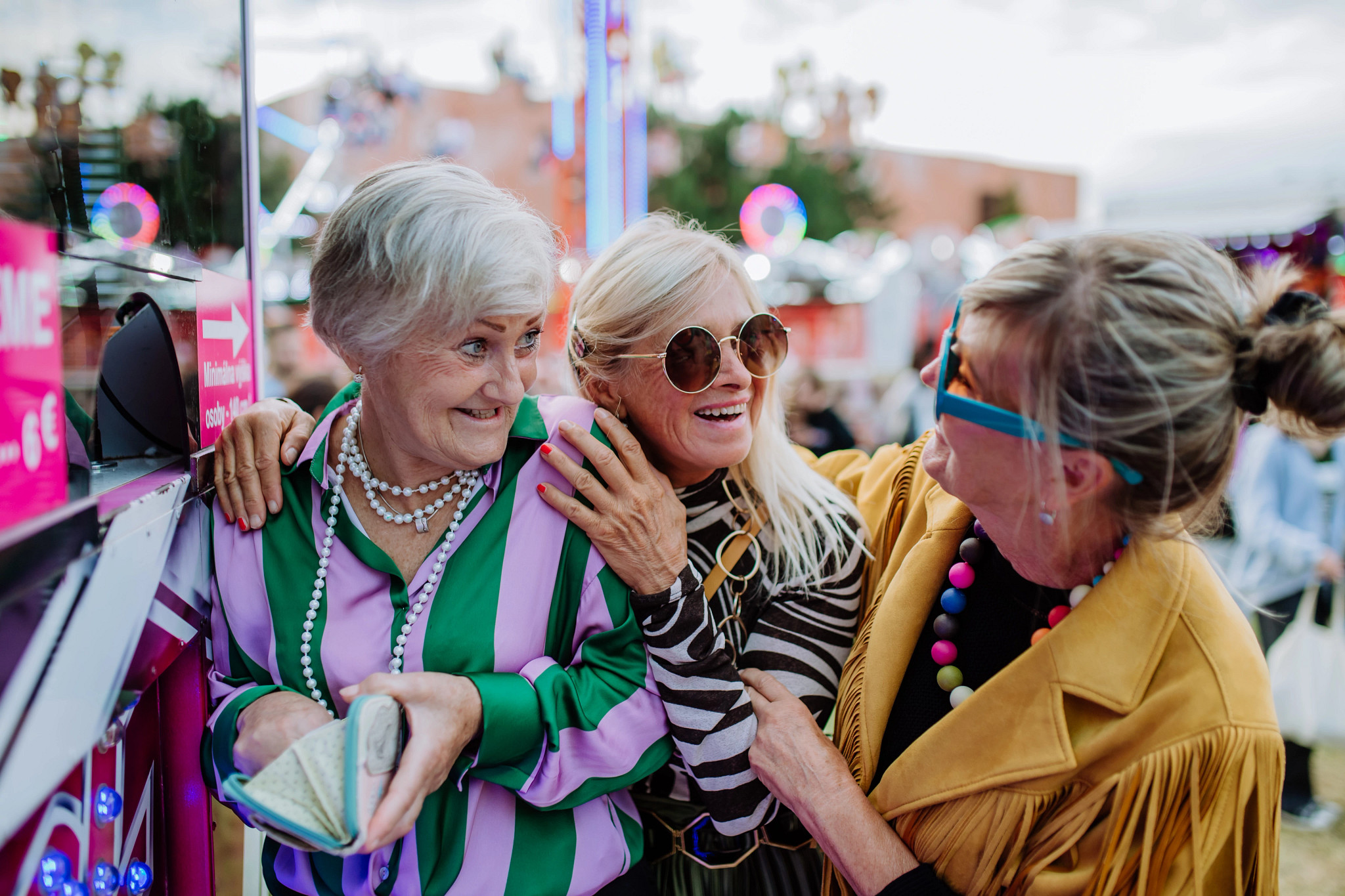 Happy three senior friends in colourful clothes enjoying county fair, buying tickets to carousel together.