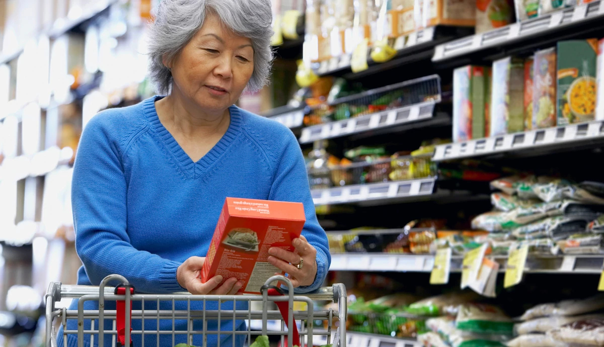 Image of an older woman, standing in a grocery store aisle, looking at an item she pulled off the shelf.