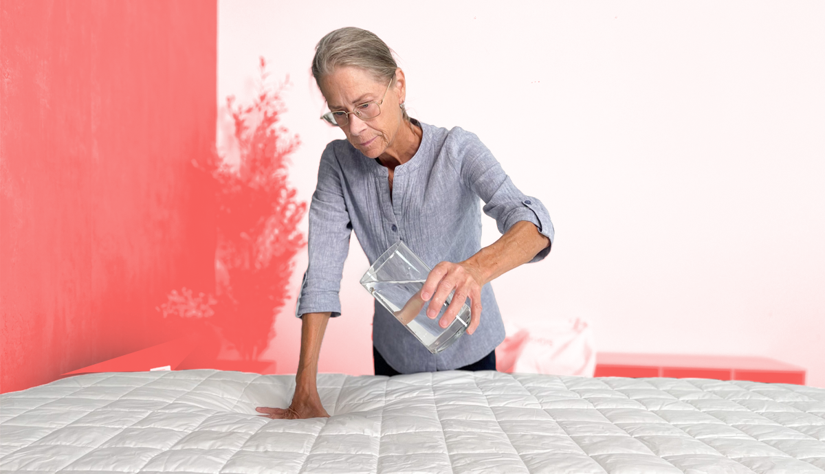 A woman pouring water from a glass onto the Saatva Signature Mattress Pad