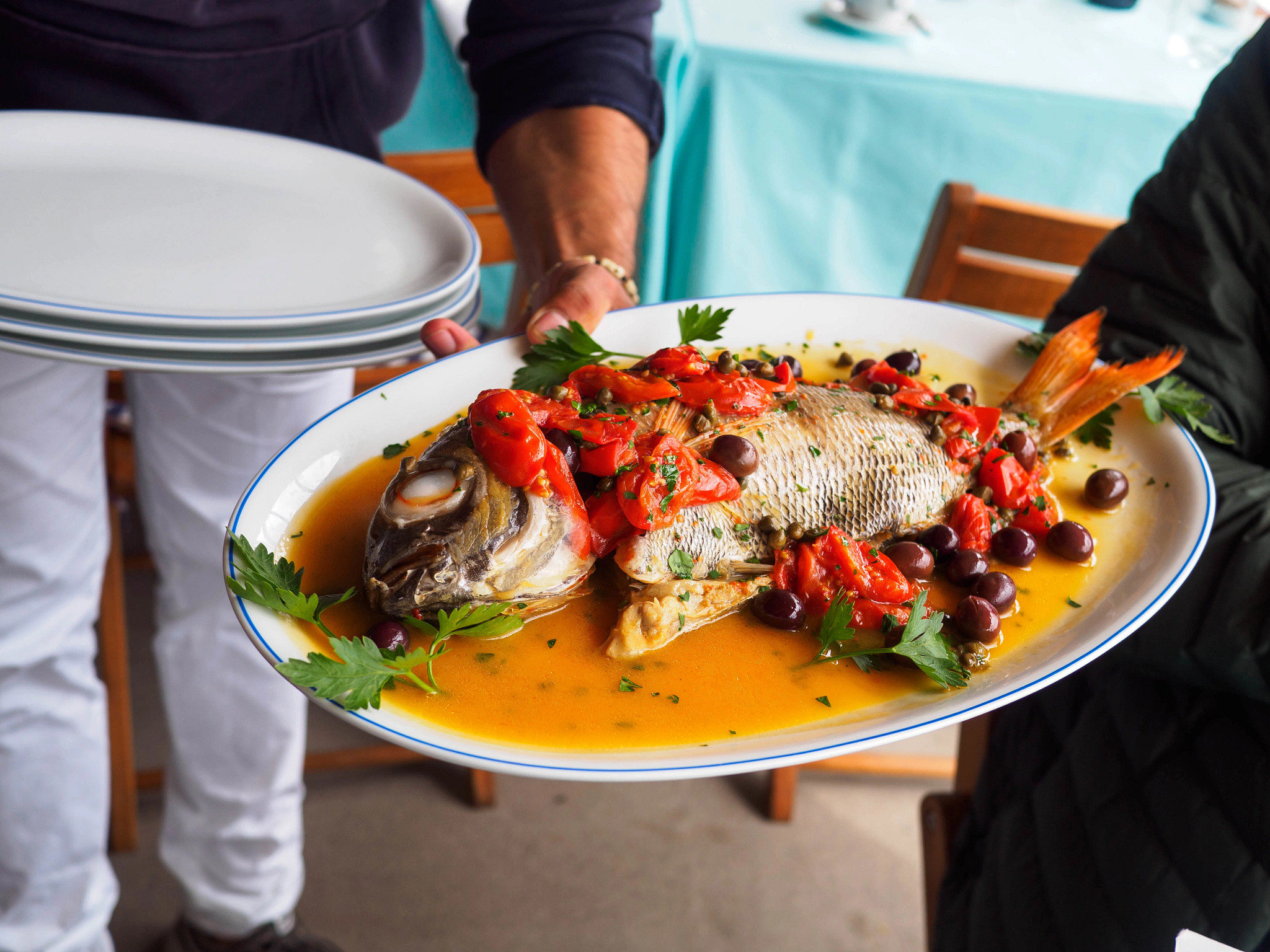 A whole fish, eyes and all, on an oval plate. The fish is topped with roasted tomatoes and kalamata olives