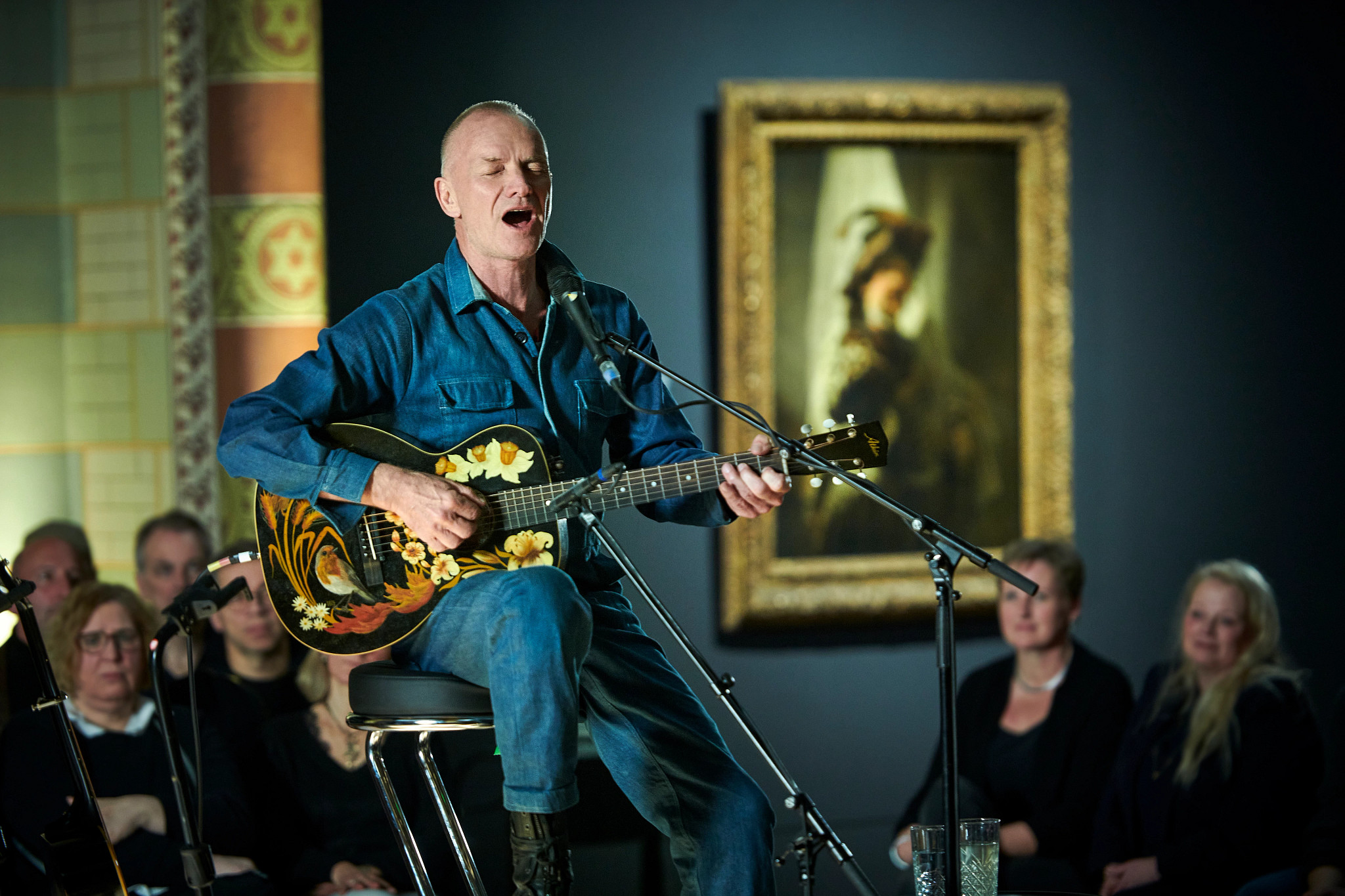 sting, seated while singing and playing a uniquely decorated guitar in front of an audience at the rijksmuseum