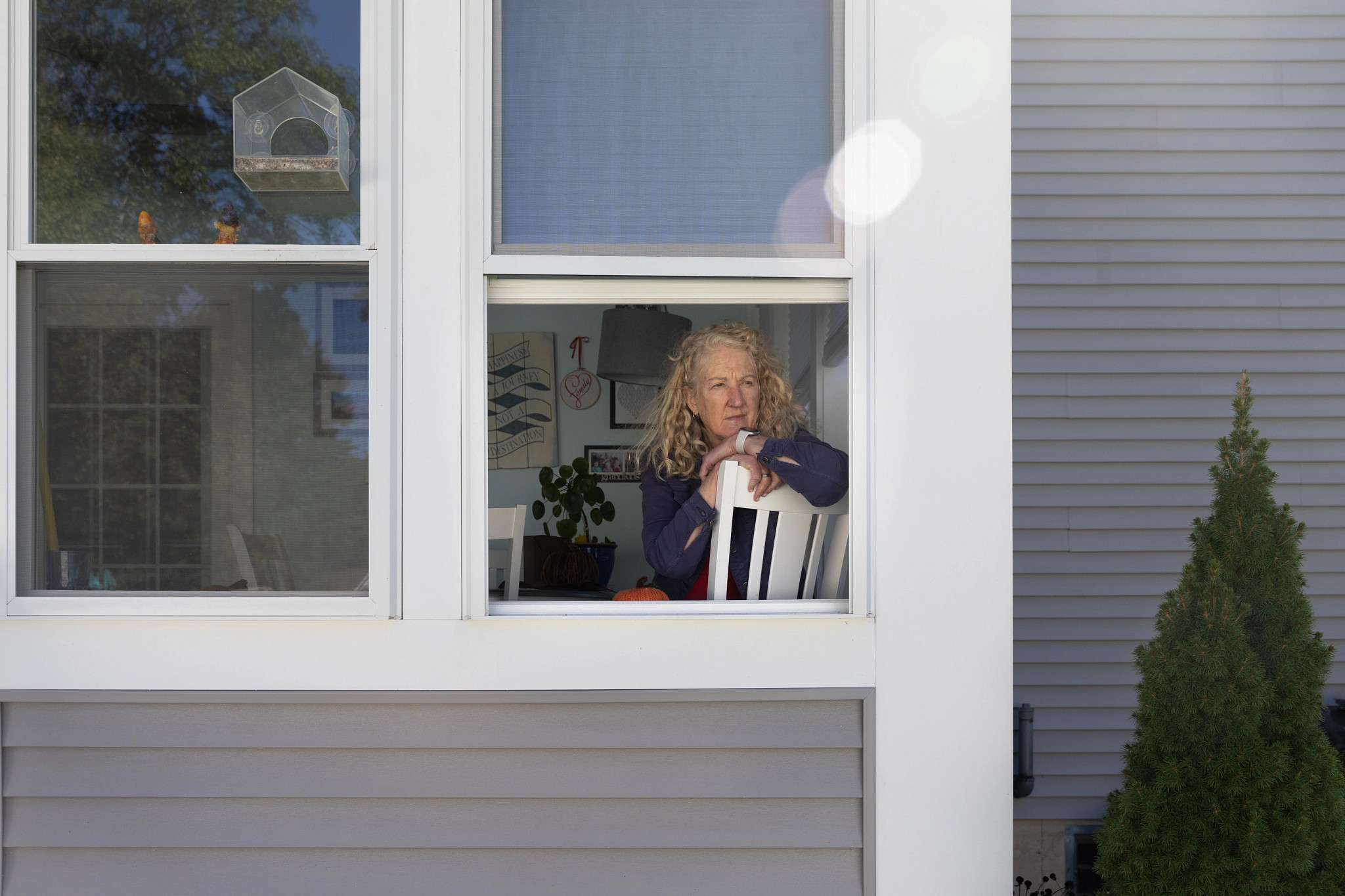 A woman leans on a white chair, looking out an open window of a house.