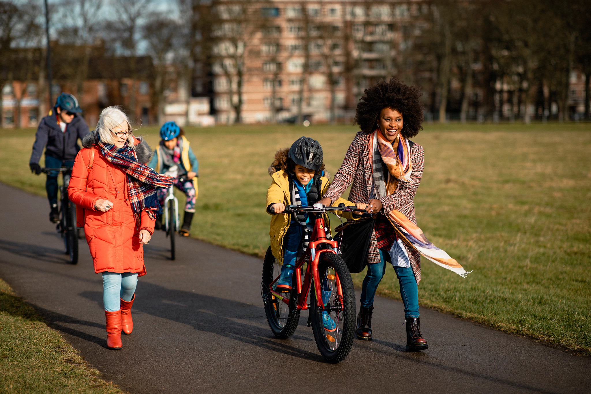 Three generation family walking through a public park. The little boy is cycling through the park with his mothers assistance while she talks too her mother in-law