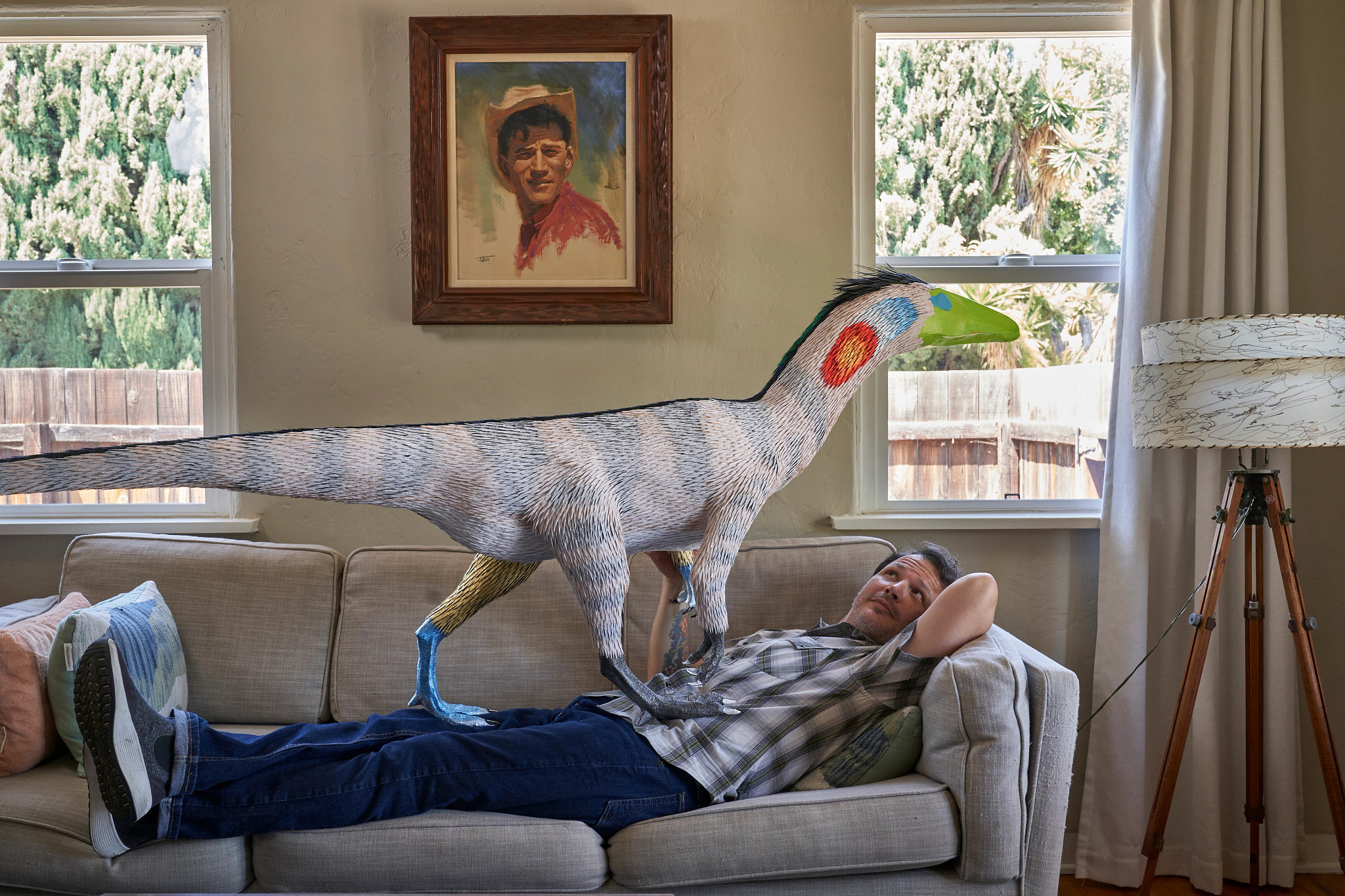 Artist Roberto Benavidez is shown in a photo lounging on his couch underneath a papier-mâché dinosaur sculpture