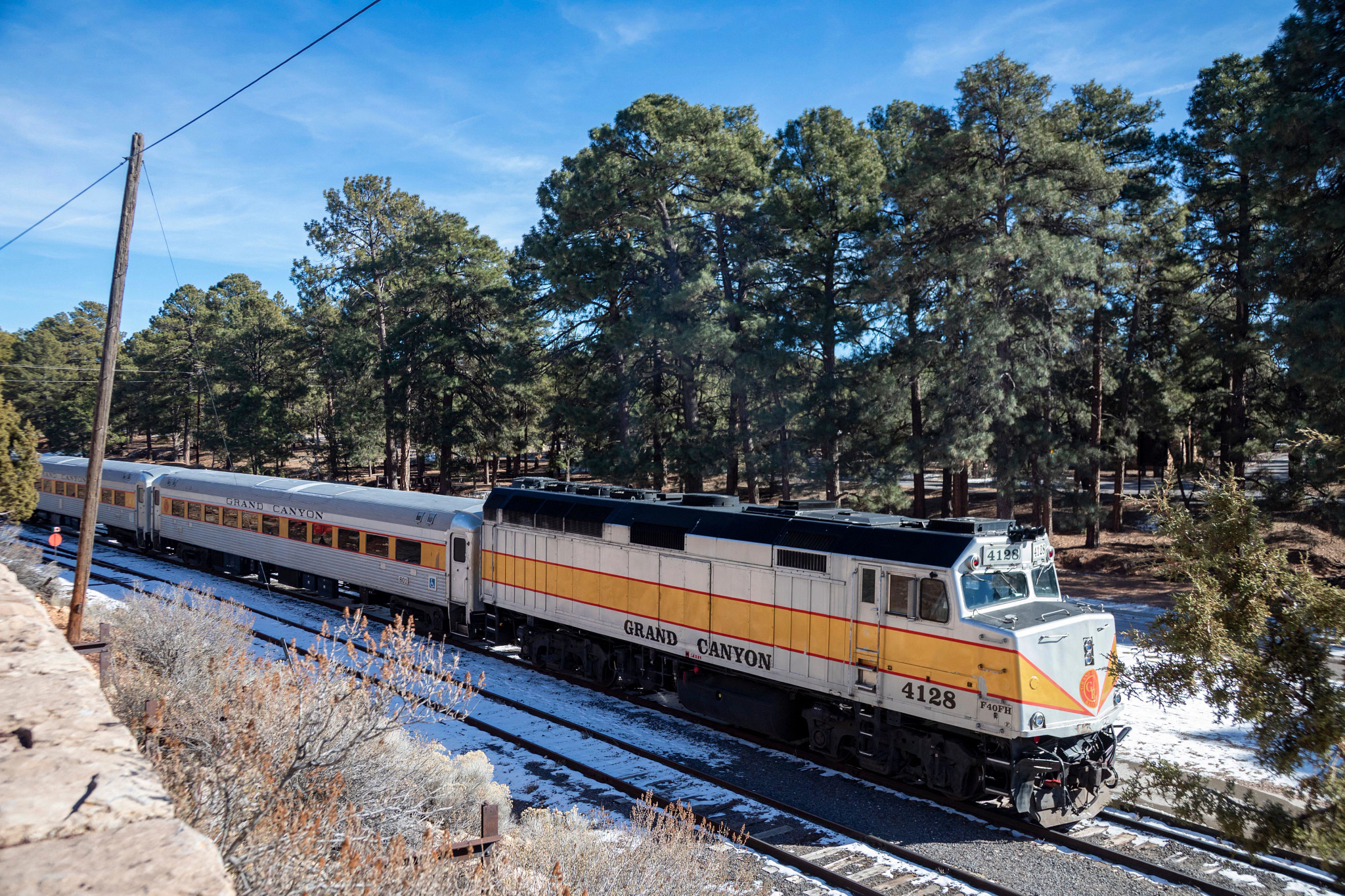 A classic Grand Canyon Railway passenger train travels through a snow-dusted pine forest under a clear blue sky