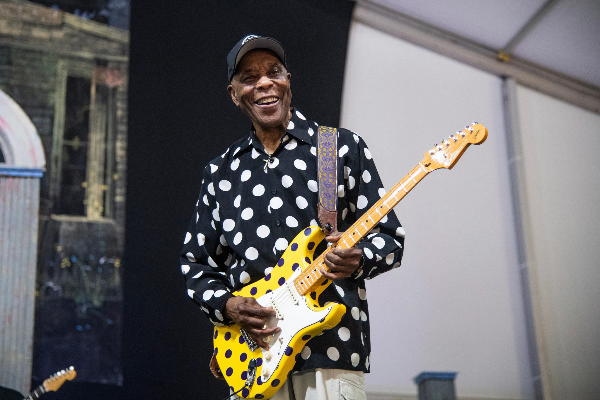buddy guy smiling, holding a yellow and purple polka dot guitar, wearing a black and white polka dot shirt and baseball cap