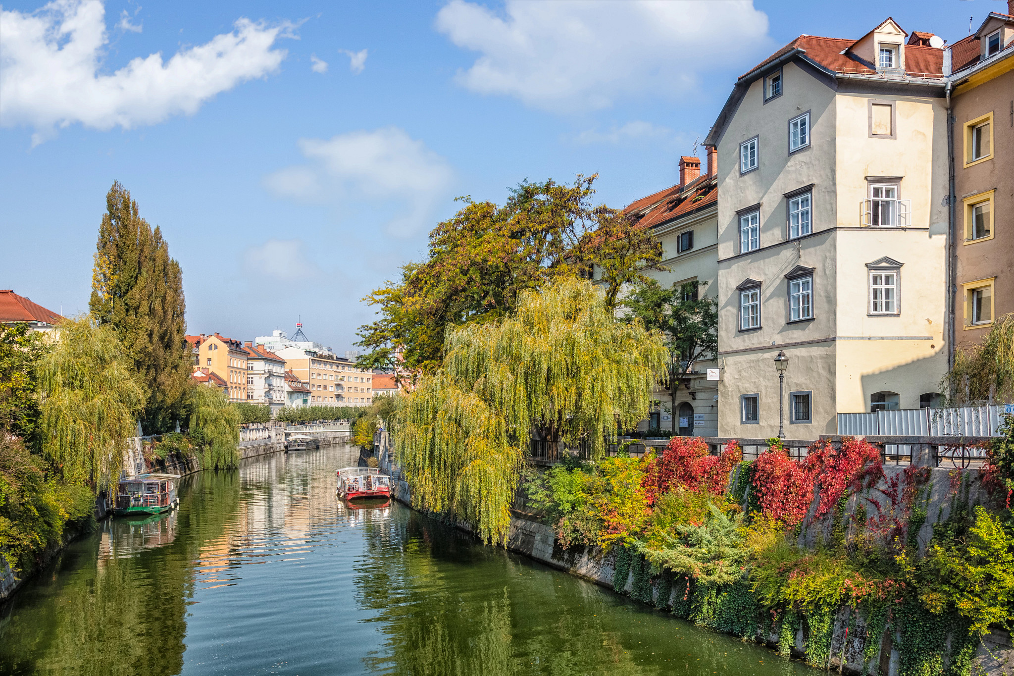 Tree-lined river flowing through a historic European neighborhood with colorful buildings, boats and autumn foliage.