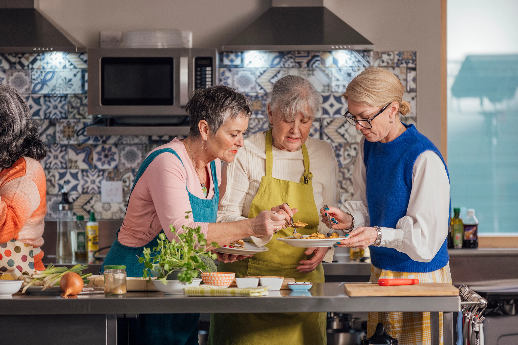 Three women stand around a stainless steel kitchen island preparing food together in a modern kitchen with a tiled backsplash.