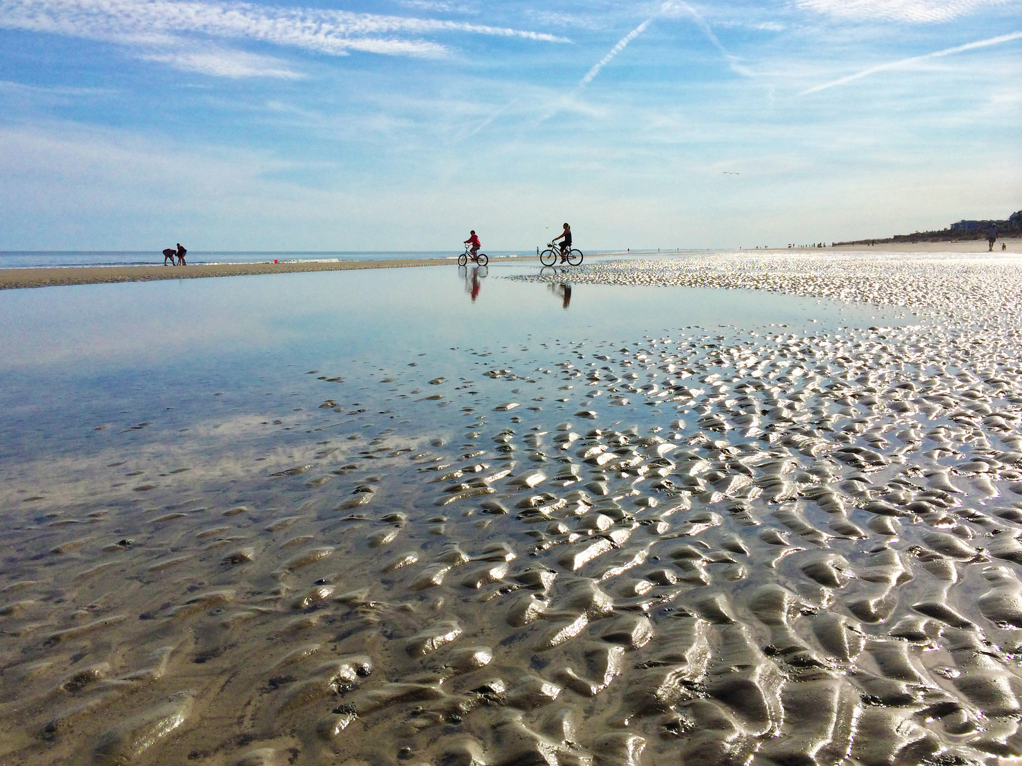 Cyclists riding near the beach shoreline during low tide, with reflective water, open sky and expansive coastal views.