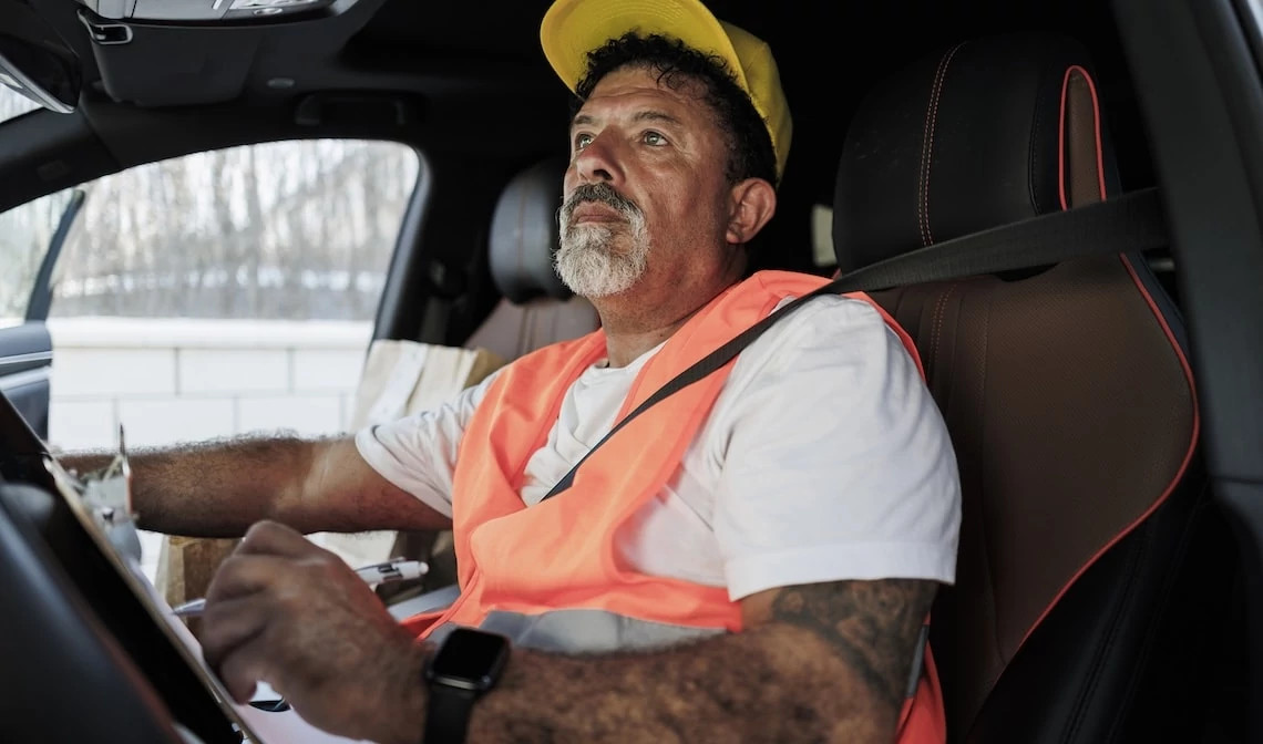 Older worker in a safety vest and hard hat preparing for work from the driver’s seat of a vehicle