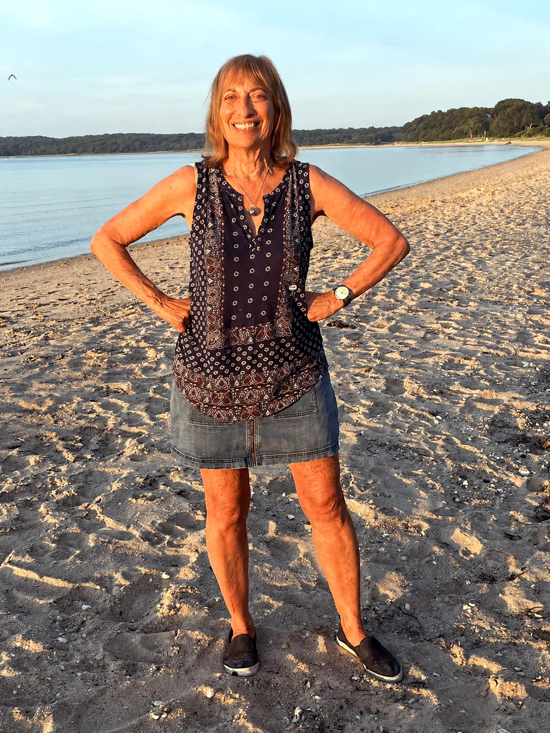 Catherine Hiller smiles on a sandy beach at sunset while standing with her hands on her hips.