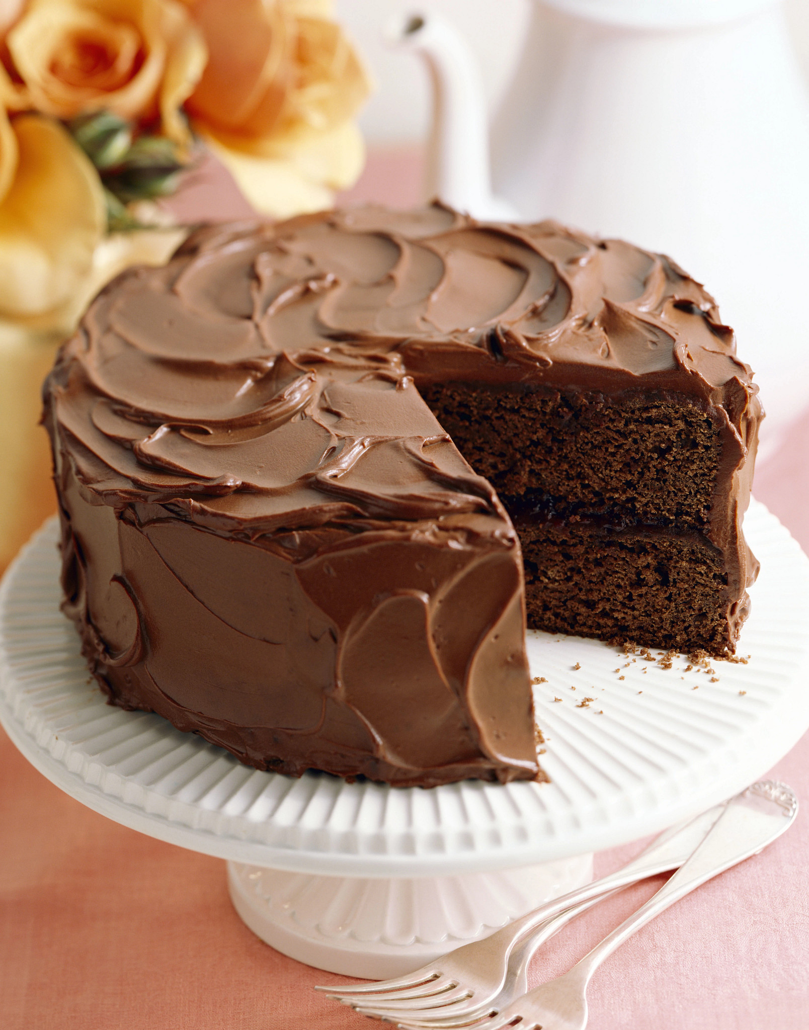Chocolate layer cake covered in thick chocolate frosting displayed on a white cake stand with a slice removed, revealing moist cake layers. Yellow roses and serving utensils appear in the background