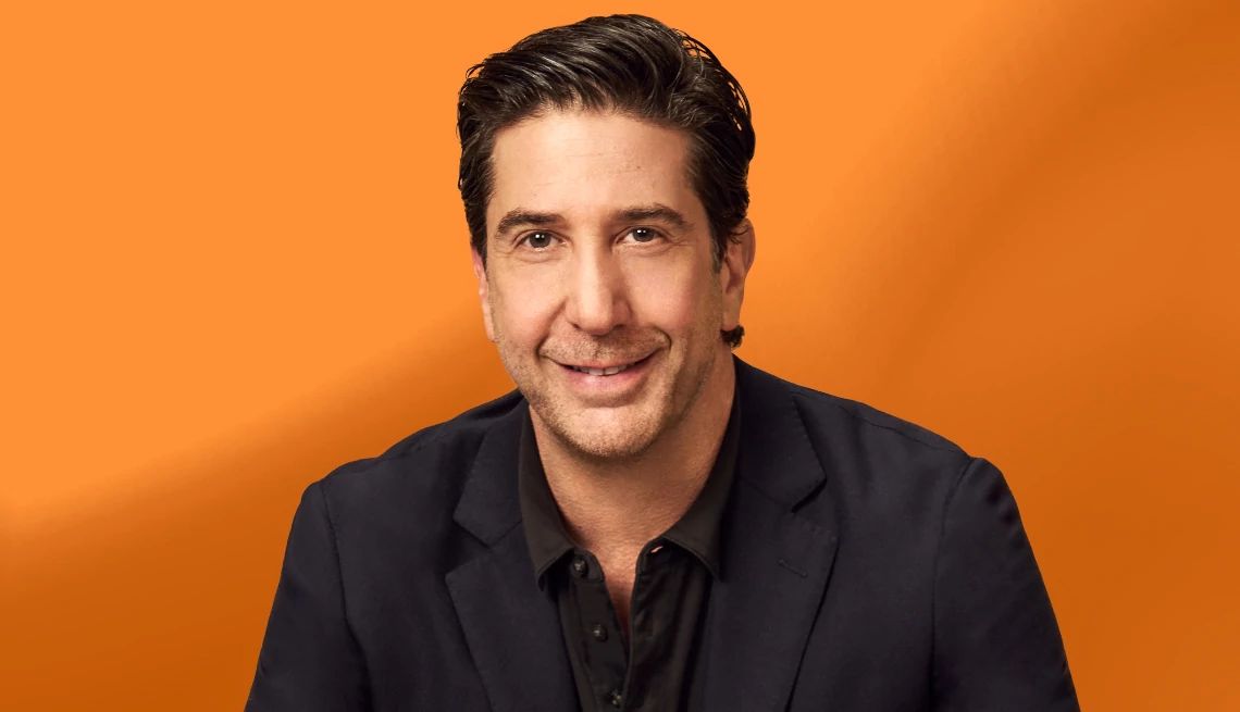 A Portrait of David Schwimme David Schwimmer in black suit, warm smile, and a backdrop of vibrant orange