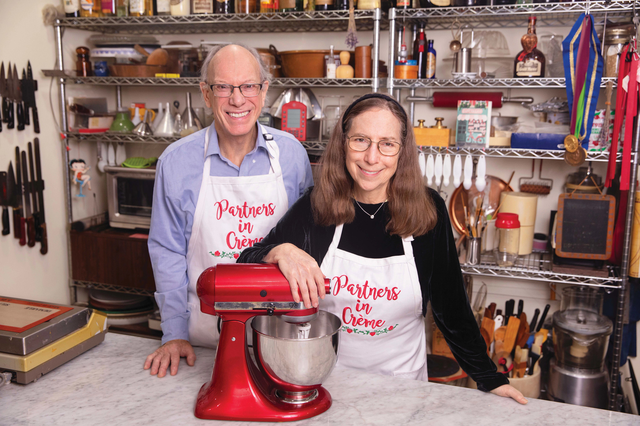 Woody Wolston and Rose Levy Beranbaum wearing aprons that say Partners in Creme with mixer on table in front of them, kitchen supplies behind them