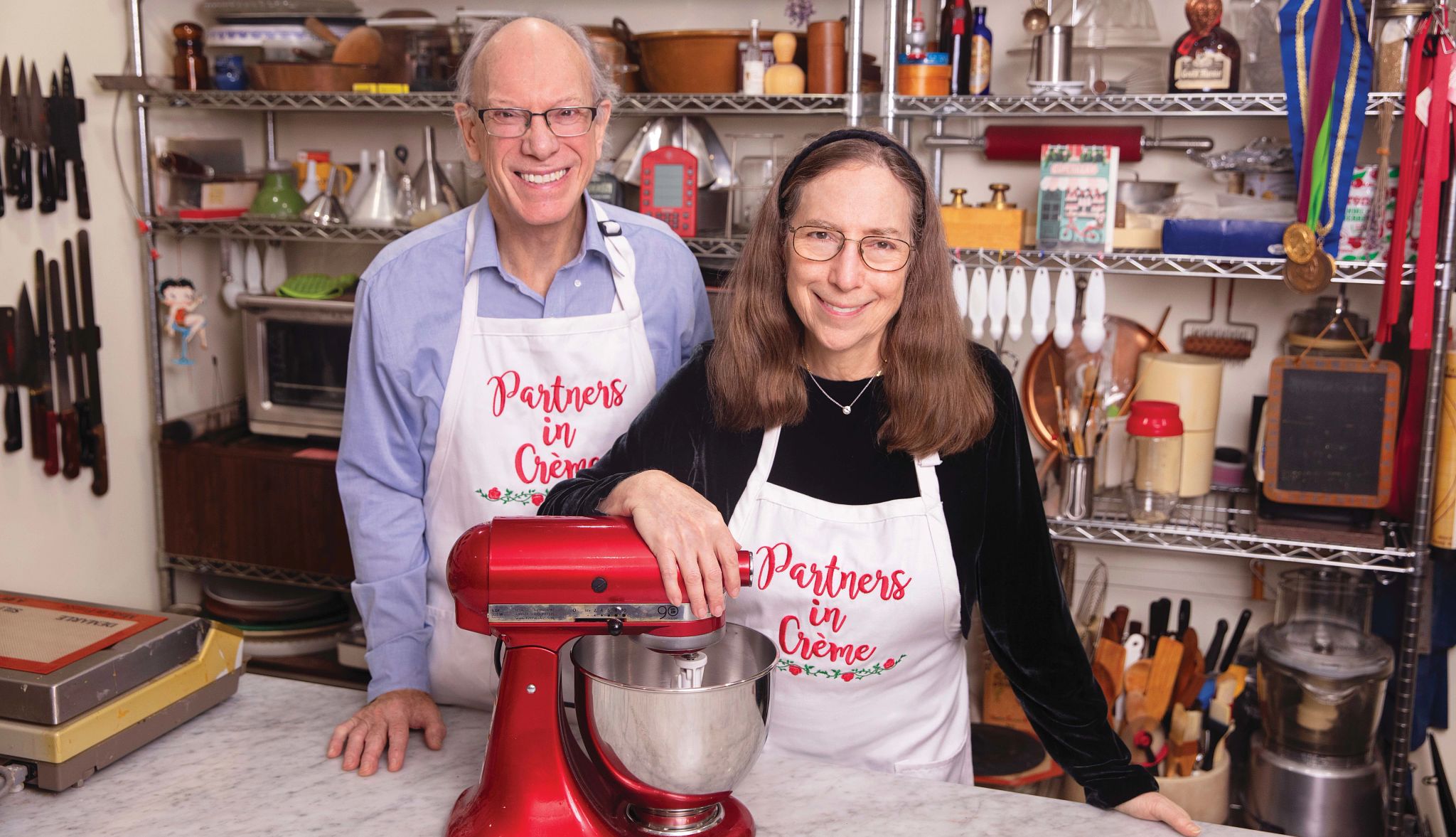 Rose Levy Beranbaum with husband Woody Wolston Woody Wolston and Rose Levy Beranbaum wearing aprons that say Partners in Creme with mixer on table in front of them, kitchen supplies behind them