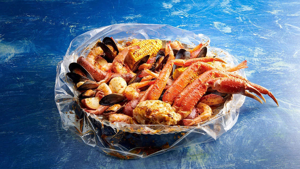 seafood boil in plastic bag on blue table and includes crab legs, corn, clams, shrimp, sausage, potatoes