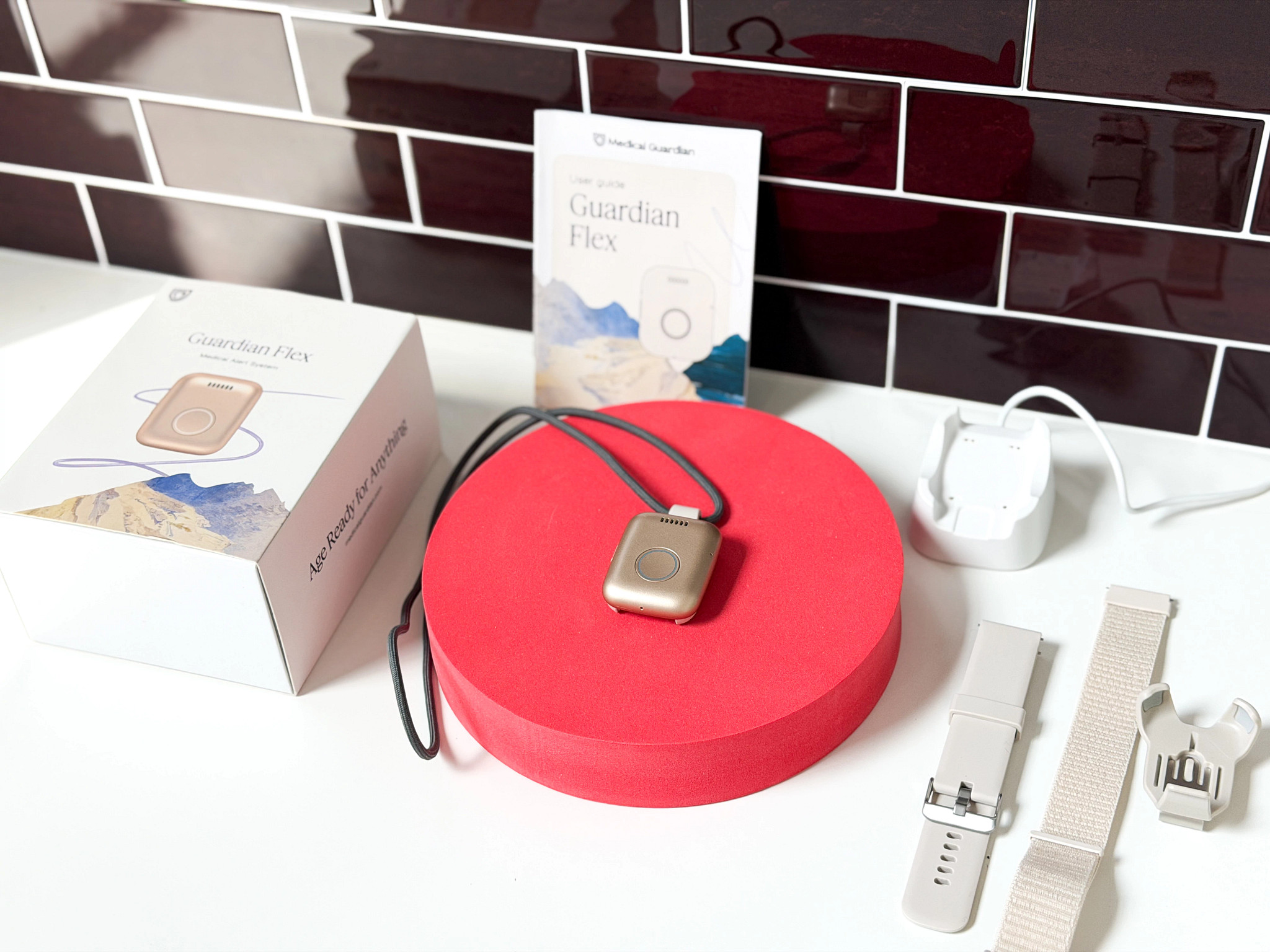 The Guardian Flex wearable medical alert system with strap from Medical Guardian displayed on a red stand with the packaging and accessories around it.