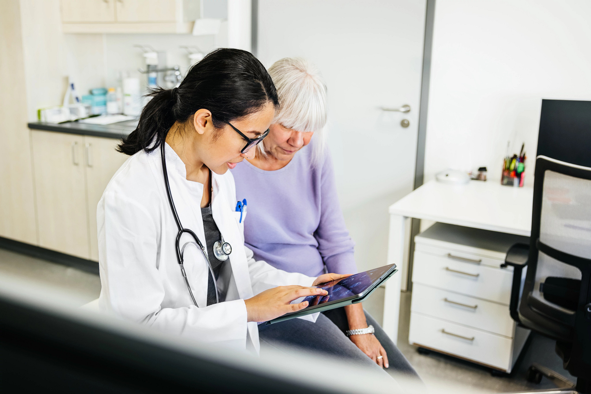 A young female doctor in a lab coat and glasses sits beside a senior patient in a modern medical office, reviewing medical information on a digital tablet. The doctor points to the screen, indicating a collaborative approach to discussing medical details or treatment plans.