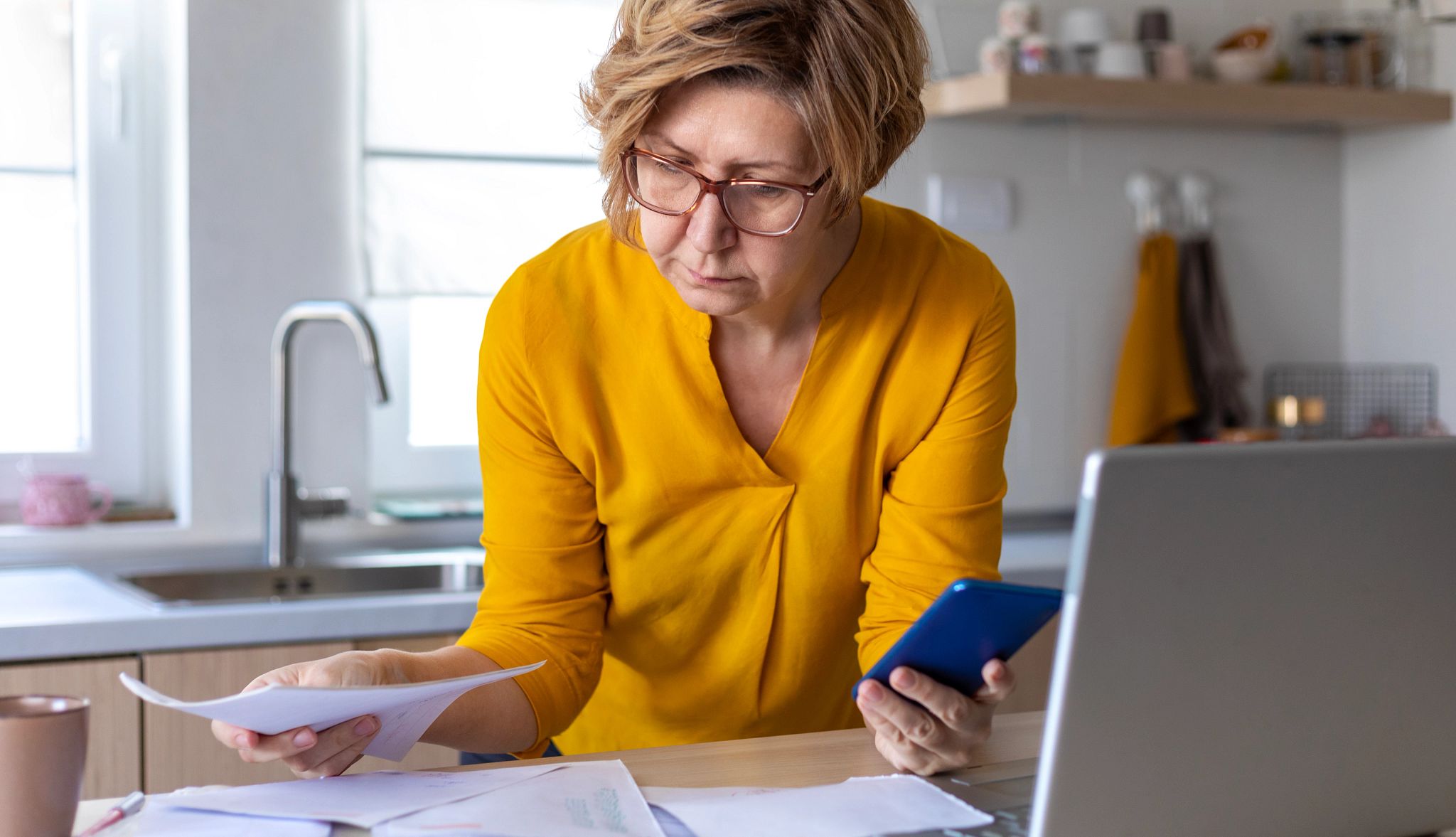 Concentrated mature woman calculating finance, money, using laptop computer and smart phone at home workplace table, counting budget, paying bills, taxes, rent, mortgage fees Concentrated mature woman calculating finance, money, using laptop computer and smart phone at home workplace table, counting budget, paying bills, taxes, rent, mortgage fees