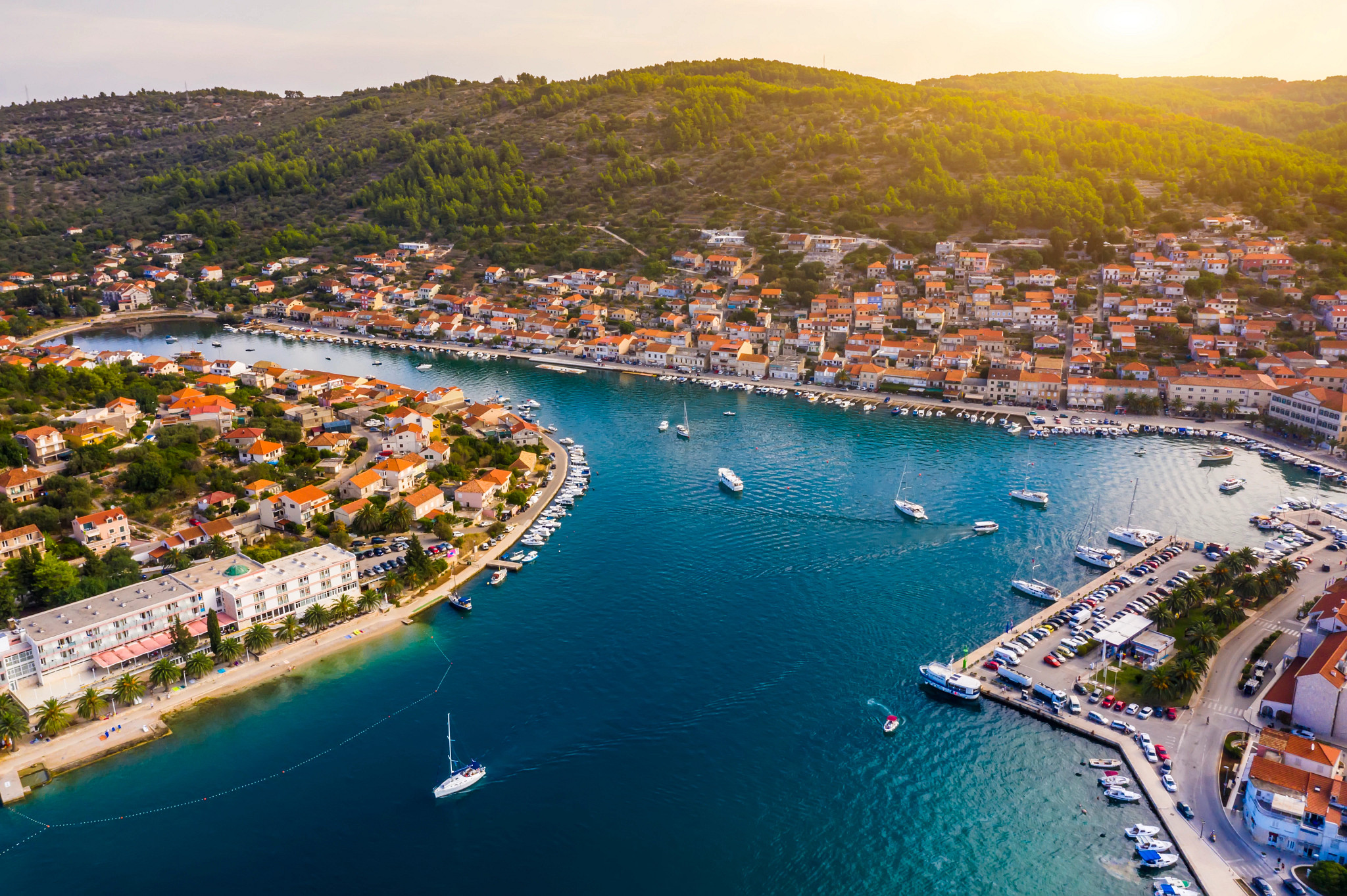 A picturesque coastal harbor town with turquoise water, sailboats, waterfront homes and green hills at sunset.