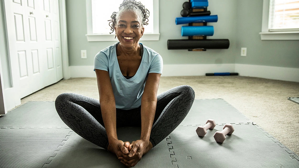 middle age woman smiling wearing workout clothes sitting with crossed legs on mat, hand weights next to her, other workout gear in background