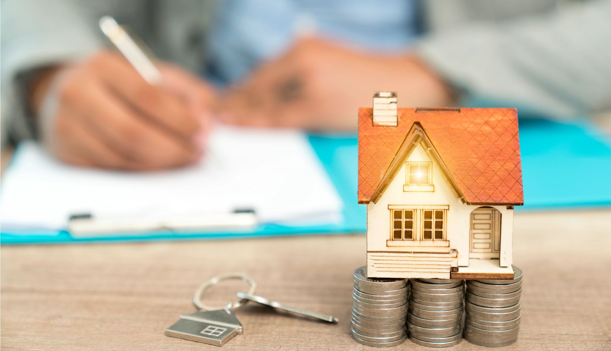 States Ban Unfair Real Estate Agreements A person signs a real estate contract behind an image of a key and a house on top of stacked coins