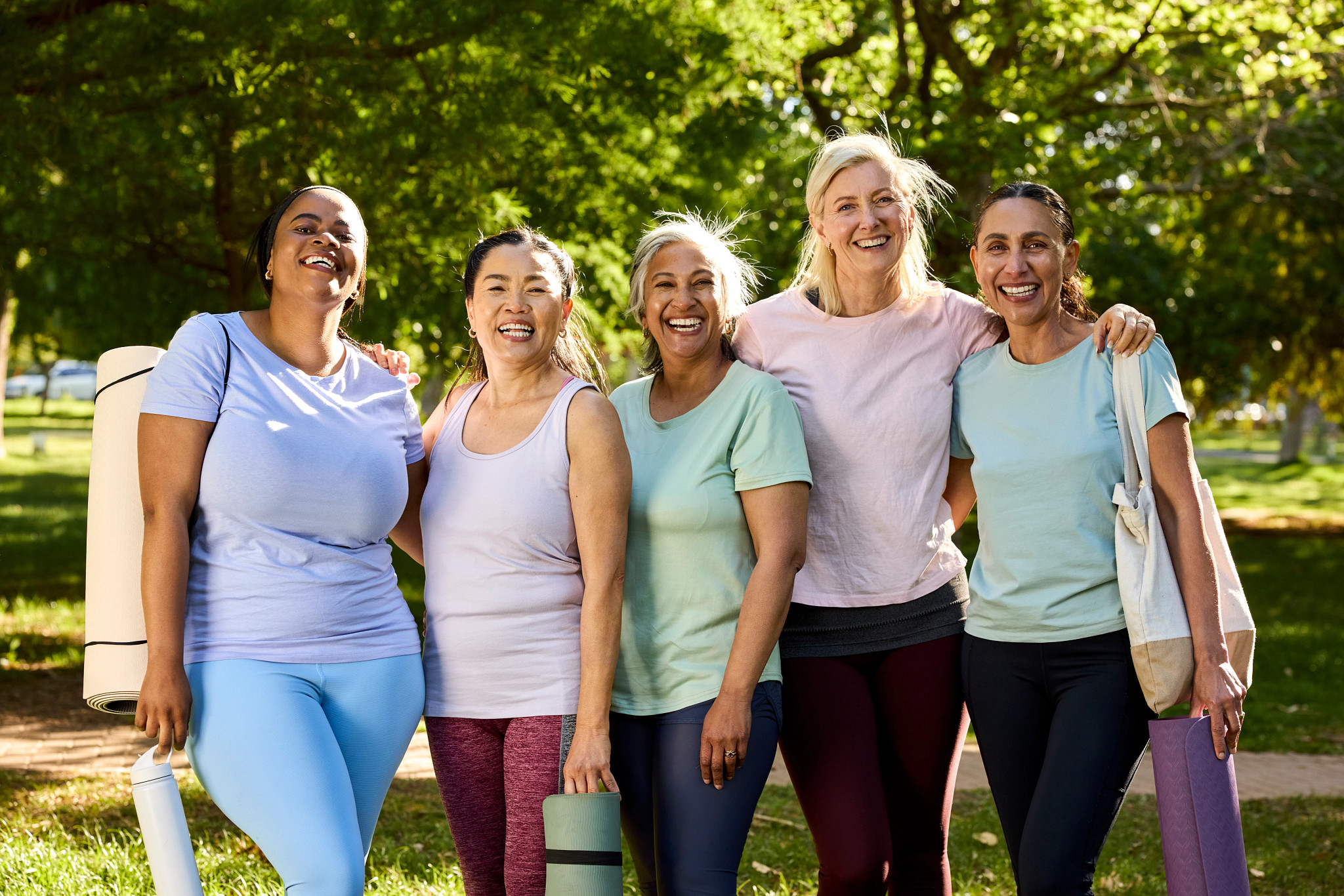 A group of five smiling women standing side by side together in a park with yoga mats