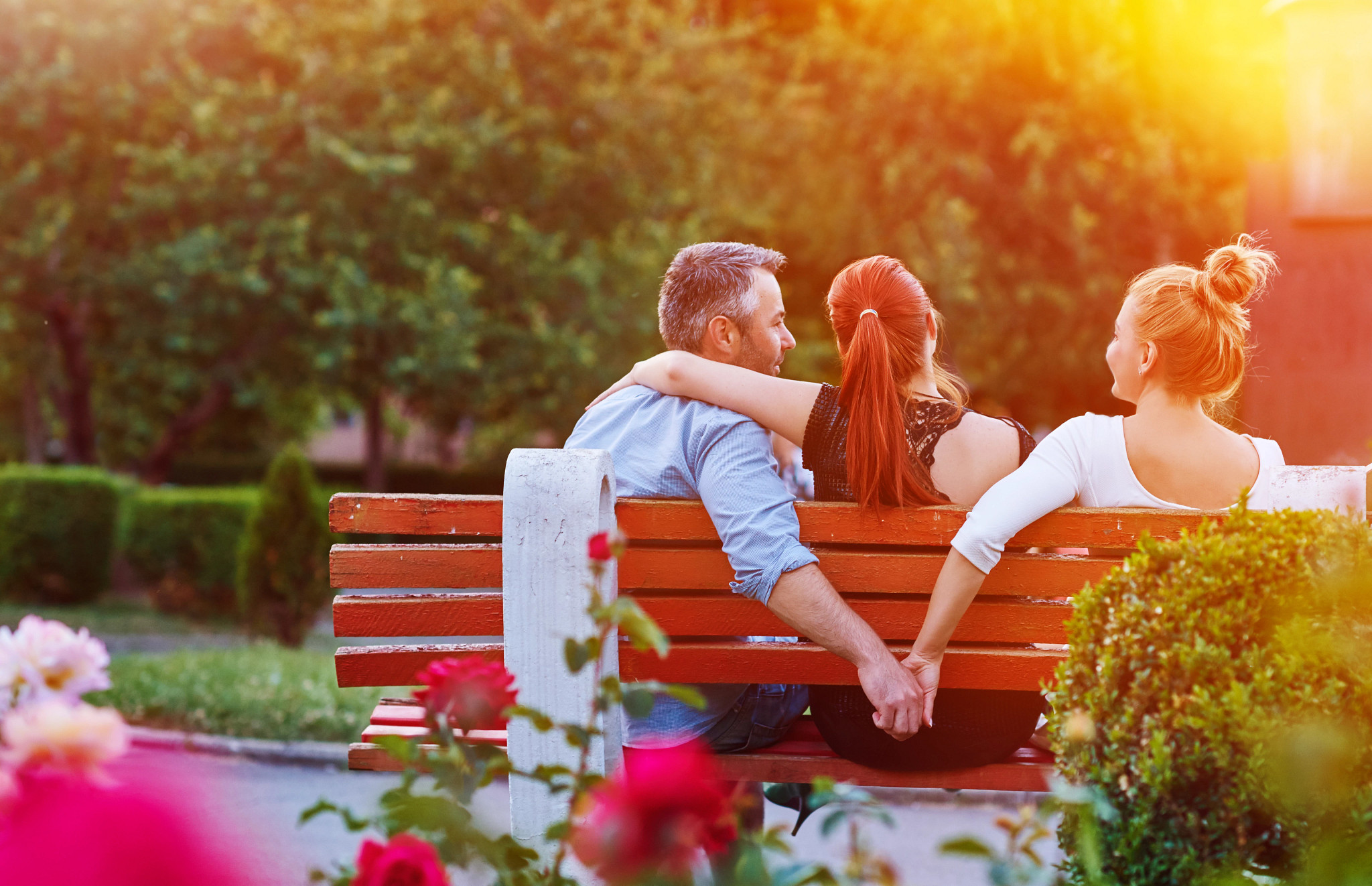 A photo of a husband and wife sitting with a woman on a park bench. While the wife has her arm around her husband's shoulders, he is holding hands with the other woman behind her back.