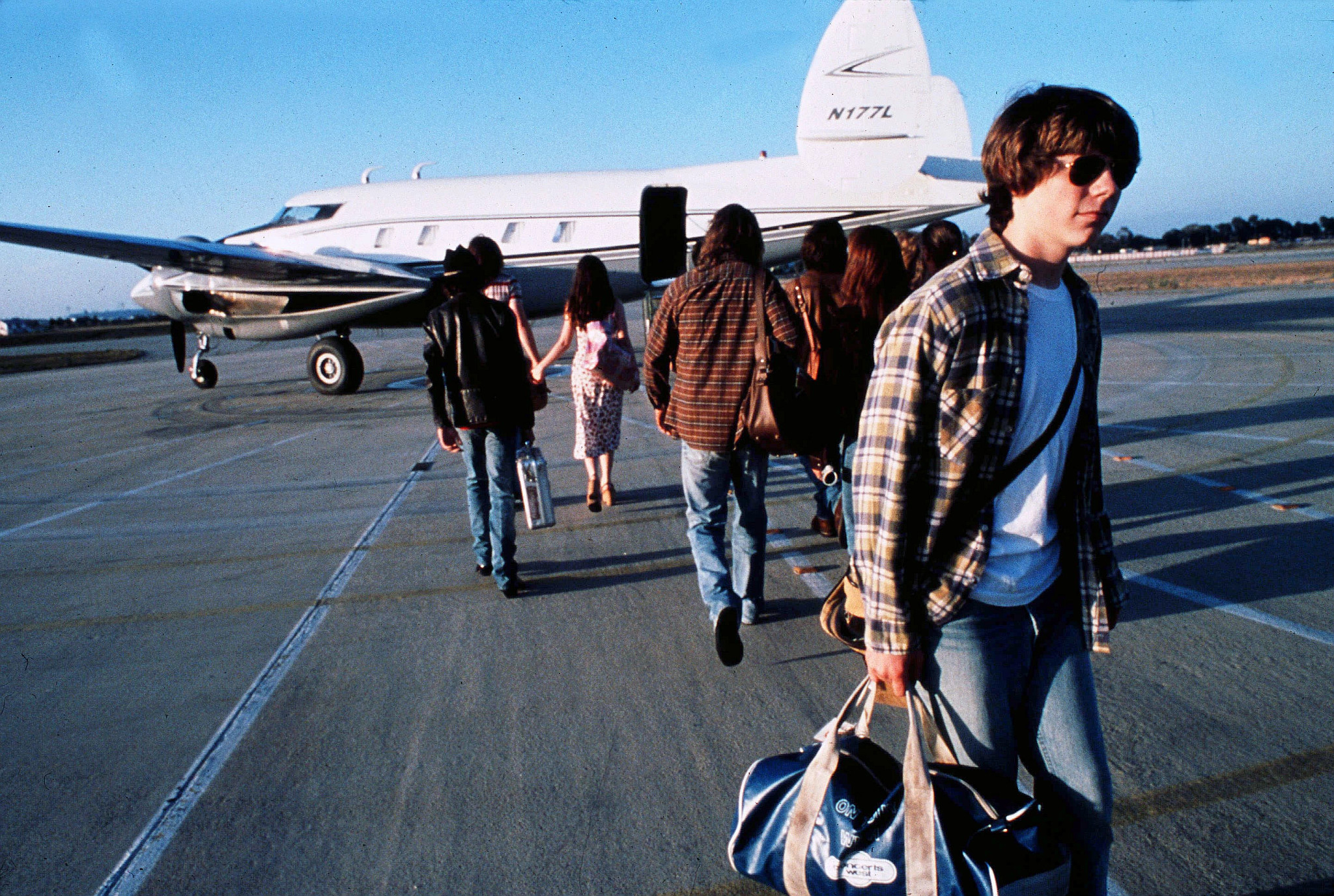Cast members of Almost Famous carrying bags walk across an airport tarmac toward a parked private jet under a clear blue sky