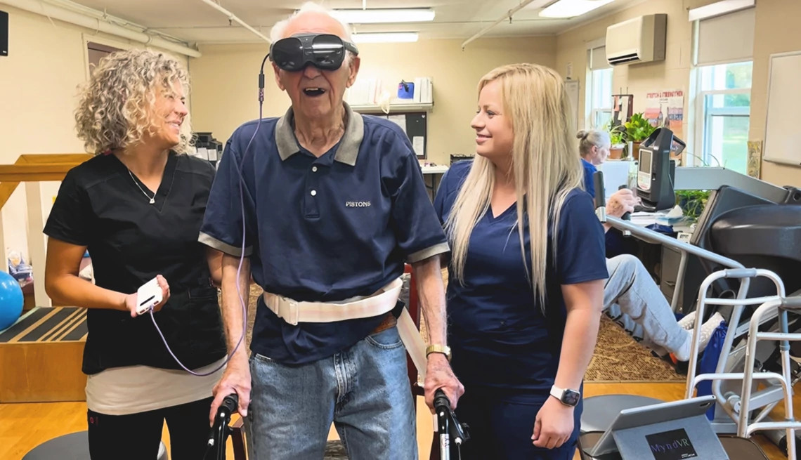 an older adult, using a walker to move, wears a virtual reality headset. he stands between two caregivers