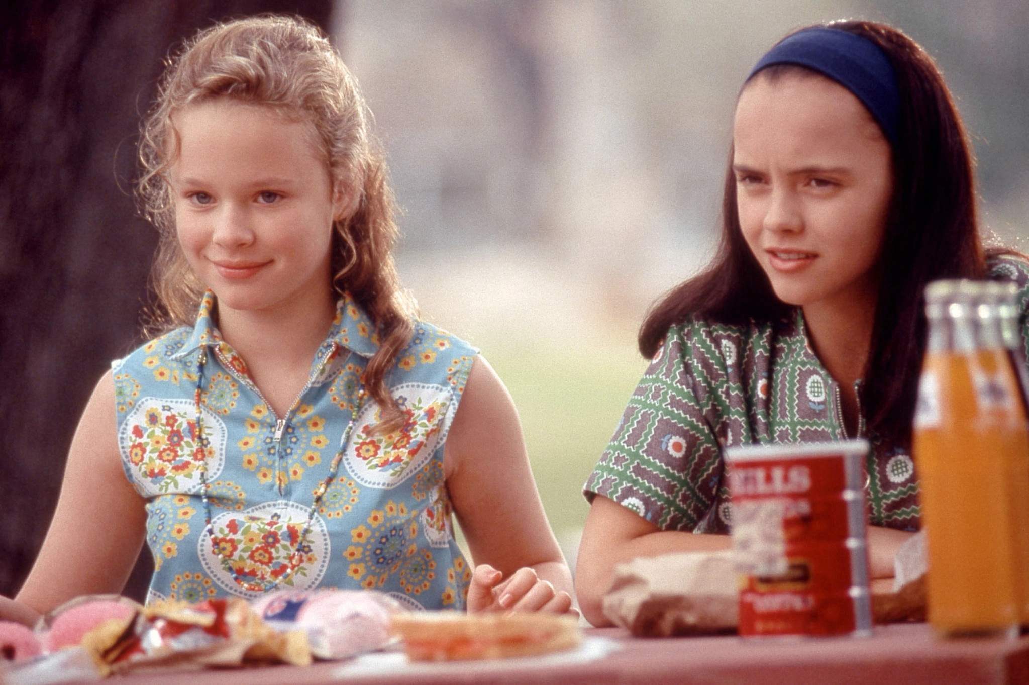 Thora Birch and Christina Ricci seated outdoors at a picnic-style table with snacks, bread, and bottled drinks in a soft warm-toned scene in the film Now and Then