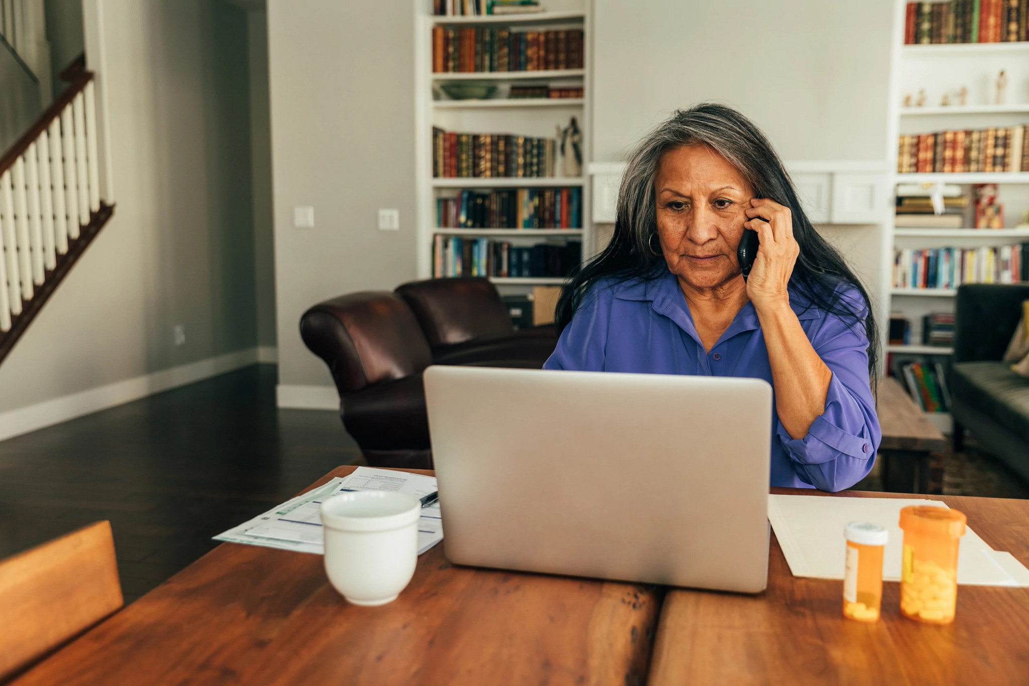 A senior aged woman sits at her kitchen table while paying medical bills, talking with her doctor, and updating medicine prescriptions.