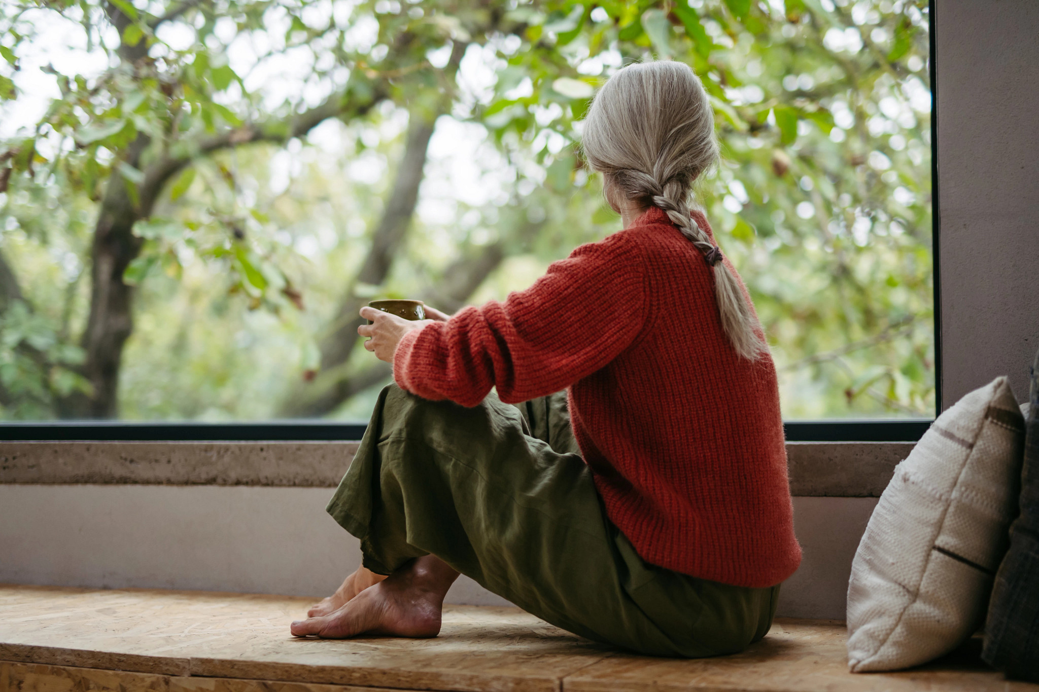 A woman sitting by a window with a coffee mug in her hand and looking outside at trees