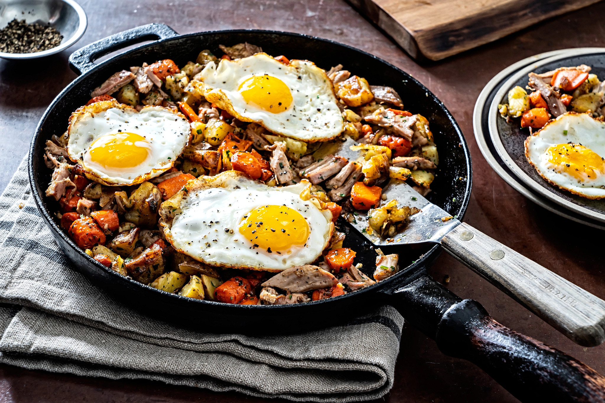 a photo shows leftover turkey hash in a frying pan, sitting on atop kitchen towels on a table. Sunny side up eggs are featured in the dish