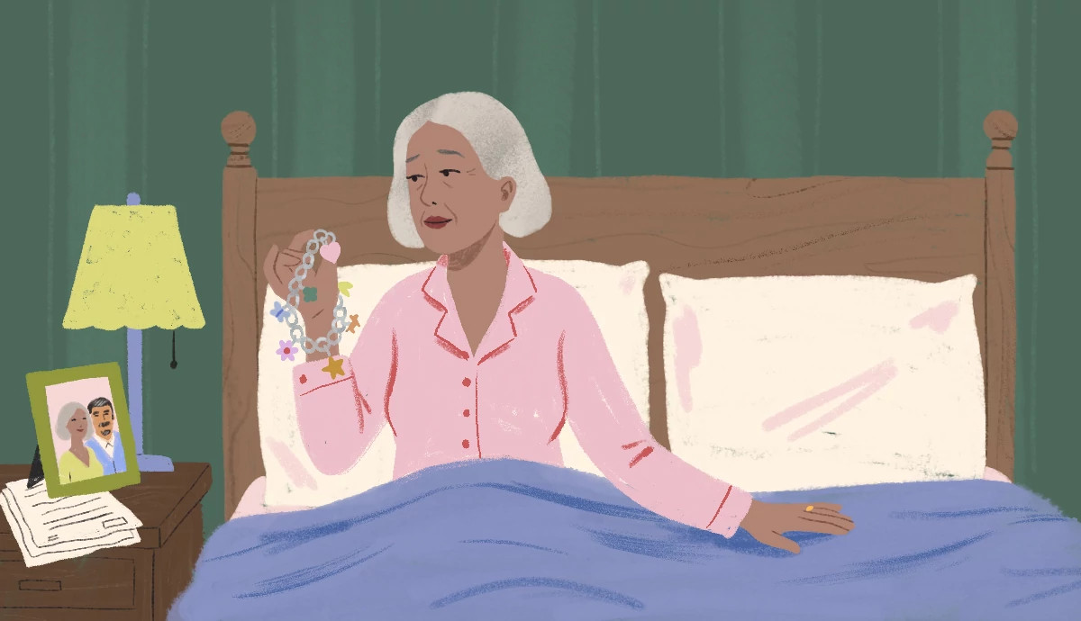 a woman sits alone in a bed, looking at a charm bracelet. on the bedside table is a photo of her with her husband.