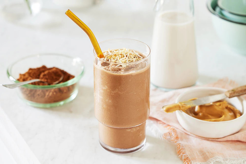 Chocolate smoothie topped with coconut flakes and served with a gold straw alongside cocoa powder, milk, and honey ingredients