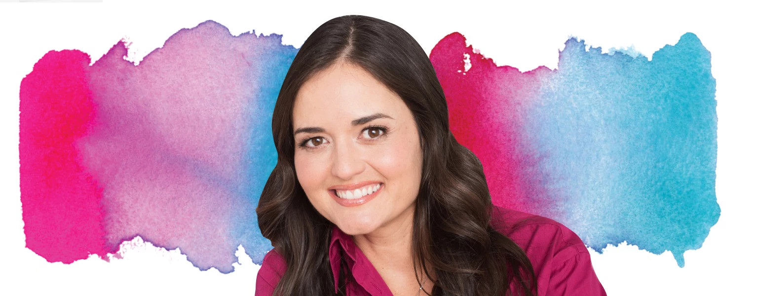 Close-up studio portrait of Danica McKellar wearing a magenta top, positioned in front of a colorful watercolor-style backdrop blending blue, pink, and purple tones