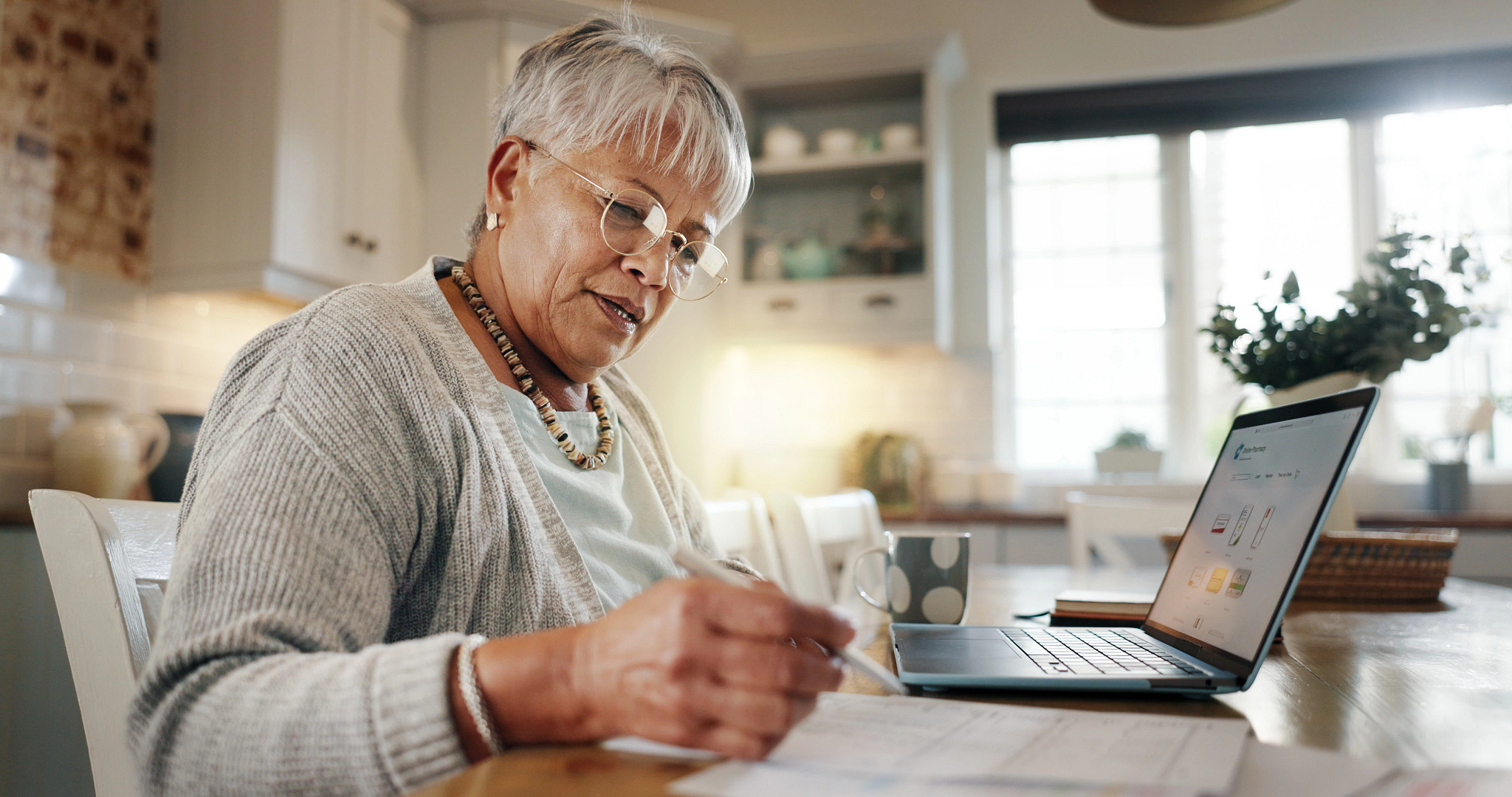 a photo shows an older adult woman working on a laptop in her kitchen