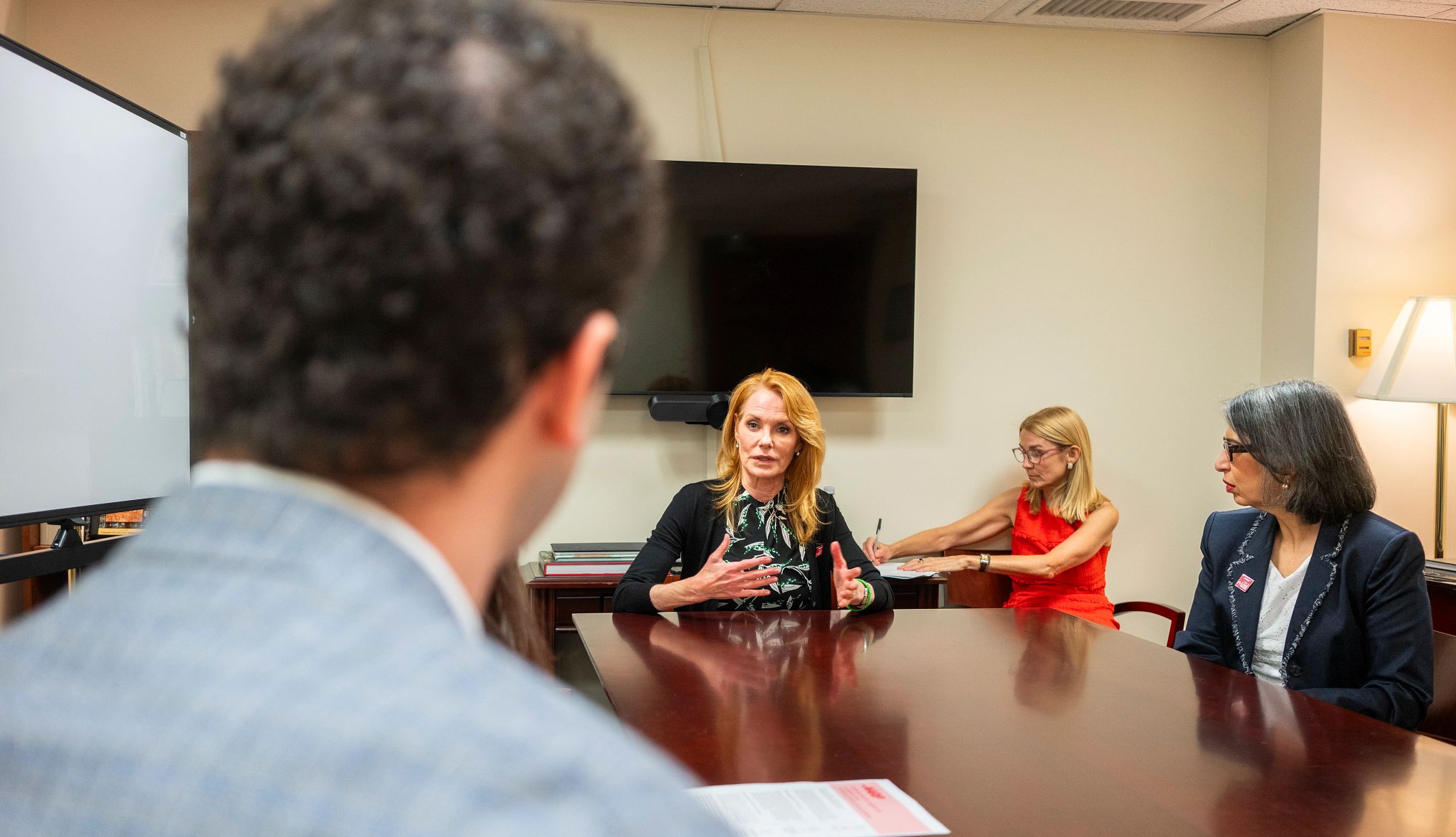 Marg Helgenberger, in a printed black dress and black sweater, speaks while sitting at a conference table.